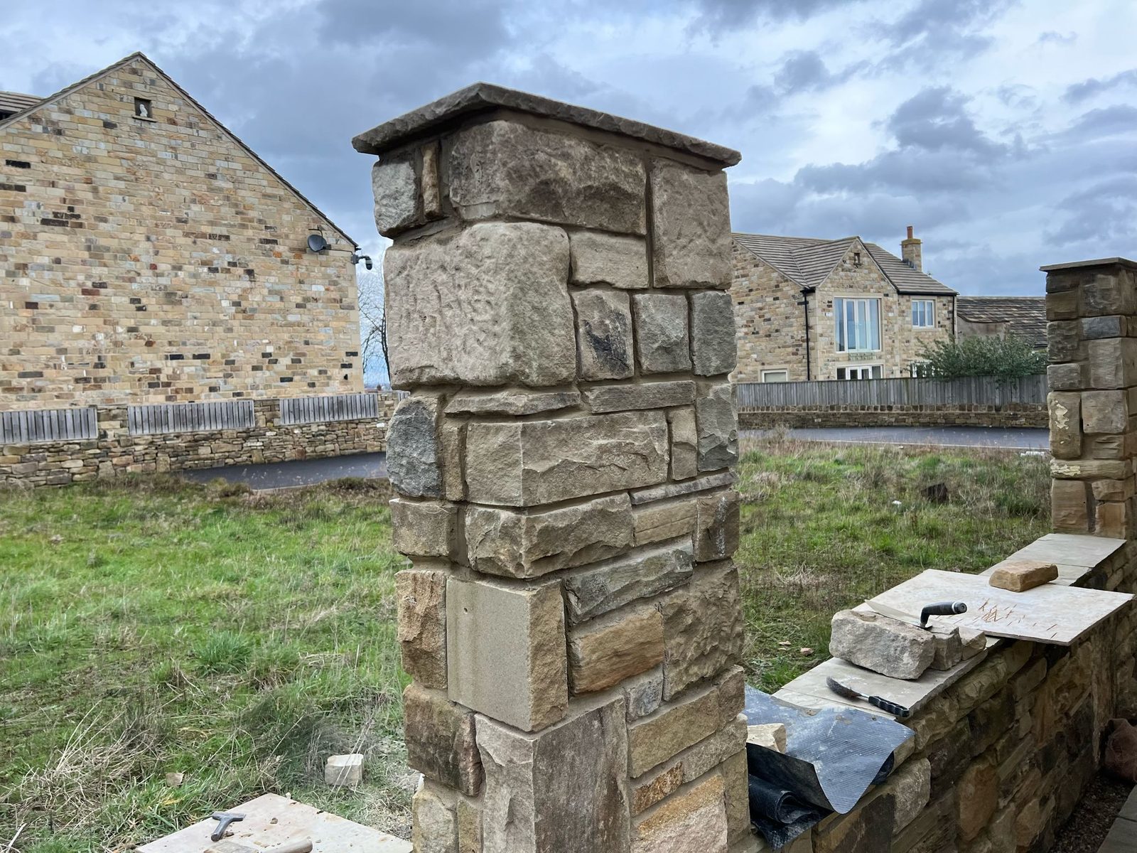 A stone gate pillar and stone wall being built, with stone houses behind